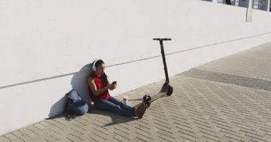 Kulaklık takan Afrikalı Amerikalı kadın akıllı telefon kullanarak kaldırımda scooterlı oturuyor.