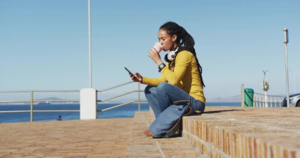 Africano americano mulher sentada usando smartphone beber café no passeio marítimo — Vídeo de Stock
