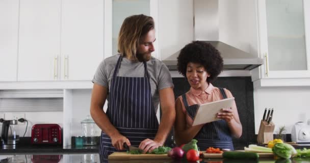 Gemischtes Paar trägt Schürzen mit digitalem Tablet und hackt gemeinsam Gemüse in der Küche — Stockvideo