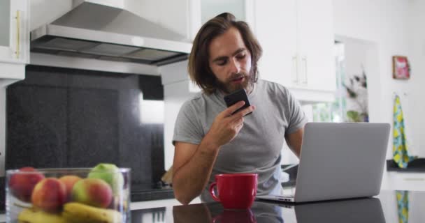 自宅で働いている間 白人男性はスマートフォンで話し キッチンでラップトップを使用しています 隔離されたまま家にいると — ストック動画