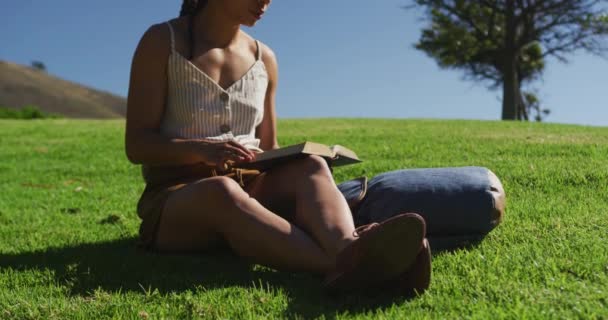 Afrykańska Amerykanka siedzi na trawie czytając książkę w parku — Wideo stockowe