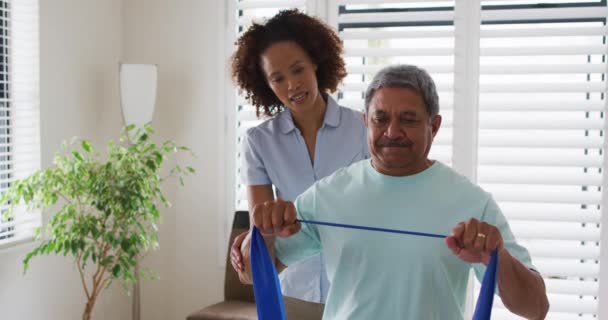 Fisioterapeuta femenina de raza mixta que ayuda al hombre mayor a hacer ejercicio con banda de ejercicio — Vídeos de Stock