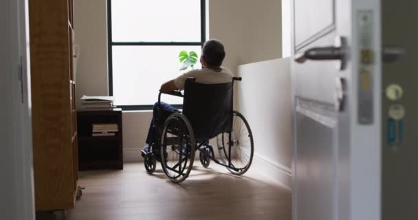 Senior mixed race man in wheelchair wearing face mask and looking through window — Stock Video