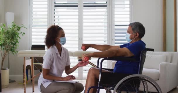 Physiothérapeute féminine de race mixte portant un masque aidant les aînés à faire de l'exercice à l'aide d'haltères — Video