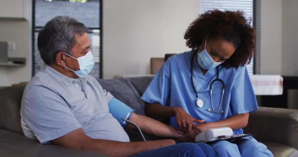 Blandad ras kvinnlig läkare bär mask och senior man som tar blodtryck hemma — Stockvideo