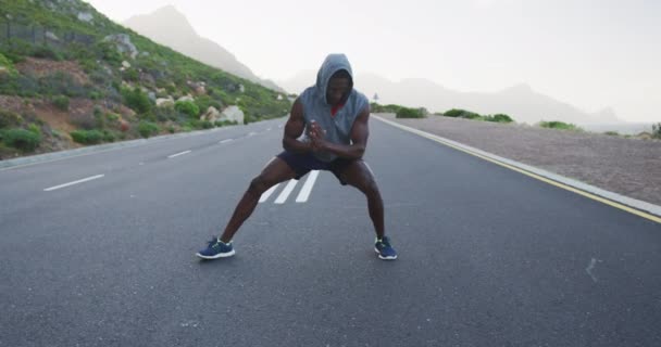 Hombre afroamericano usando una sudadera con capucha realizando ejercicio de estiramiento en el camino — Vídeos de Stock