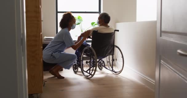 Médico femenino de raza mixta con máscara que ayuda a un hombre mayor en silla de ruedas en casa — Vídeos de Stock