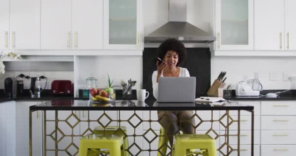 アフリカ系アメリカ人の女性が自宅で働いている間 スマートフォンで話し 台所のラップトップを使用しています 隔離されたまま家にいると — ストック動画