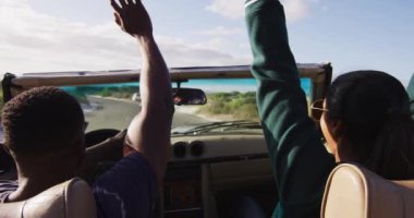 Çeşitli çiftler güneşli bir günde üstü açık bir arabada araba kullanıyor ellerini havaya kaldırıyor ve gülümsüyor. Yaz gezisi sahil kenarındaki bir kır yolunda.