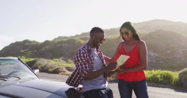Afrika kökenli Amerikalı çift, yol üzerinde üstü açık arabaların yanında dururken haritaları yol tarifi olarak kullanıyor. Yolculuk ve macera konsepti