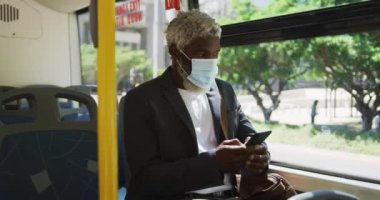 Otobüste otururken akıllı telefon kullanarak yüz maskesi takan Afrikalı Amerikalı son sınıf öğrencisi. Coronavirus covid-19 salgınında hijyen ve sosyal mesafeler.