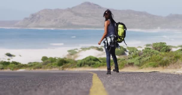 バックパックでアフリカ系アメリカ人の女性は 道路を歩いている間にリフトをヒッチしようとしています 旅と冒険のコンセプト — ストック動画