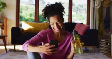 Karışık ırk kadını su içiyor ve evde yoga minderinde otururken akıllı telefon kullanıyor. Karantina karantinasında tek başına evde kalmak