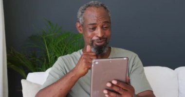 Kıdemli Afro-Amerikan adam evdeki kanepede otururken dijital tablette bir video görüşmesi yapıyor. Karantina karantinasında tek başına evde kalmak