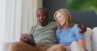 Evdeki kanepede otururken dijital tablet kullanan son sınıf melez çift. Karantina karantinasında tek başına evde kalmak