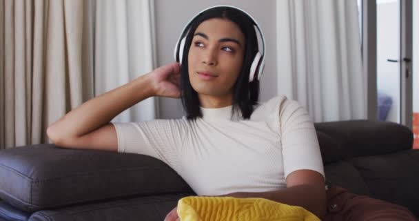 在家里坐在沙发上 性别流畅的男性戴着耳机听音乐 性别表达 特征和多样性的概念 — 图库视频影像