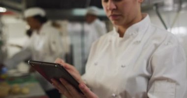 Kafkas kadın aşçı restoran mutfağında tablet kullanıyor diğer aşçılar da arka planda. Yoğun bir lokanta mutfağında çalışıyorum..