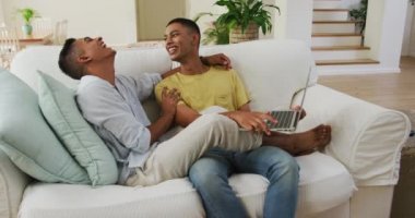 Gülen melez eşcinsel çift kanepeye oturmuş dizüstü bilgisayar kullanıyor ve konuşuyor. Karantina süresince evde tecrit altında kalmak.