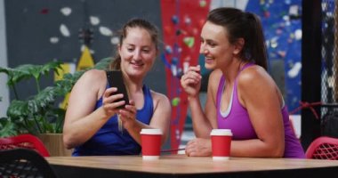 İki mutlu Kafkasyalı kadın gülüyor ve kapalı bir kafede duvara tırmanırken akıllı telefona bakıyor. fitness ve boş zaman.