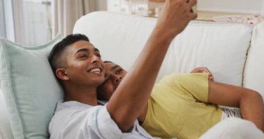 Gülen melez eşcinsel erkek çift kanepeye uzanıp selfie çekiyorlar. Karantina süresince evde tecrit altında kalmak.