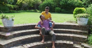 Gülen melez eşcinsel çift merdivenlerde oturmuş bahçede sarılıyorlar. Karantina süresince evde tecrit altında kalmak.