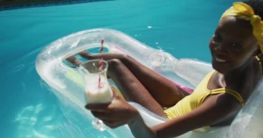 Çekici Afrikalı Amerikalı kadın şişme bir içkinin üzerinde oturuyor ve yüzme havuzunda gülümsüyor. Karantina süresince evde tecrit altında kalmak.