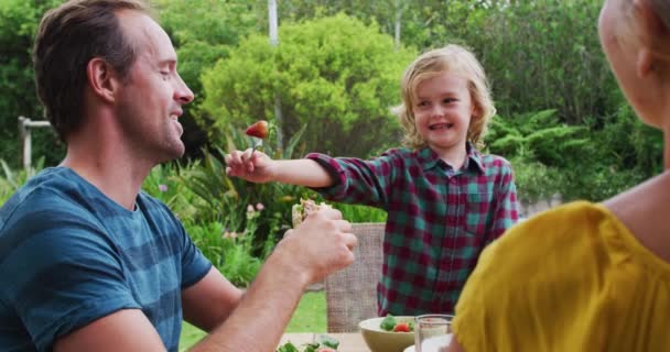 庭で家族の食事中に父親にイチゴを与える白人の少年を笑っています 一緒に屋外で食べることを祝う家族 — ストック動画