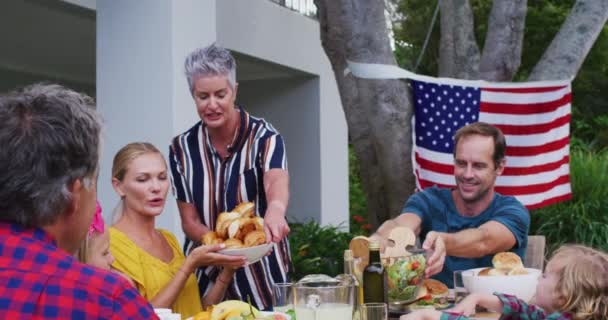 庭で一緒にお祝いの食事をする家族にサービスを提供する笑顔の白人のシニア女性 独立記念日を祝う3代目の家族が一緒に屋外で — ストック動画