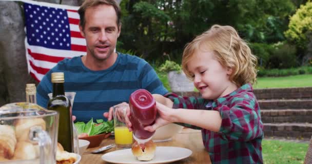 庭でのお祝いの食事中にケチャップを使って息子を見ている白人男性を笑っています 独立記念日を祝う家族みんなで屋外で食事 — ストック動画