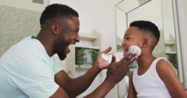 Afrikalı Amerikalı baba oğlunun ağzına tıraş kremi sürüp birlikte gülüyor. Karantina süresince evde tek başına kalmak..