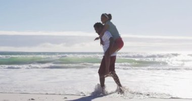 Afrika kökenli Amerikalı bir adam gülümsüyor ve Afrika kökenli Amerikalı bir kadını kumsalda sırtında taşıyor. sağlıklı açık hava eğlence zamanı deniz kenarında.