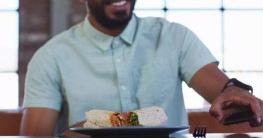 Kafede oturup, akıllı telefonuyla sandviçinin fotoğrafını çeken ve gülümseyen mutlu bir melez. dijital göçebe hareket halinde.