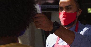 Yüz maskesi takan melez erkek kafe çalışanı kadın iş arkadaşının ateşini ölçüyor. Coronavirüs covid 19 salgını sırasında küçük bağımsız iş.
