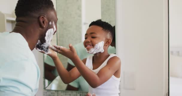 アフリカ系アメリカ人の少年は父の顔にシェービングクリームを塗り 一緒に笑う 隔離されたまま家にいると — ストック動画