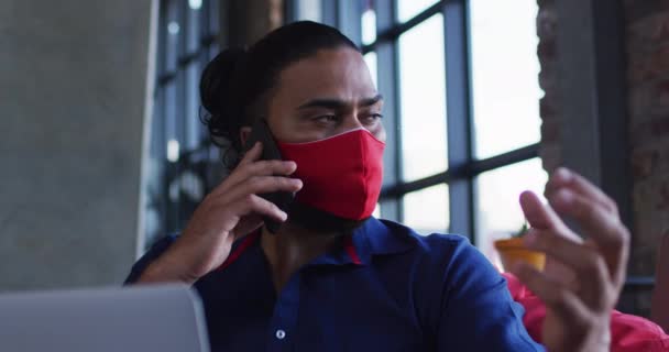 アフリカ系アメリカ人の男性は 顔のマスクをしてカフェでノートパソコンを使ってスマートフォンで話して座っている コロナウイルスの流行期の外出先でのデジタル遊牧民は — ストック動画