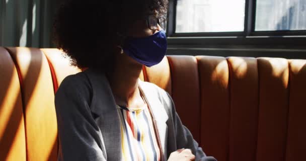 アフリカ系アメリカ人の女性が顔のマスクをしてカフェの窓を見て座っている コロナウイルスの流行期の外出先でのデジタル遊牧民は — ストック動画