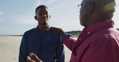 Afrikalı Amerikalı baba genç oğluyla kumsalda duruyor, elini omzuna koyup konuşuyor. Sağlıklı açık hava aile birlikte zaman geçirir..