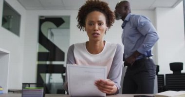 Karışık ırk işkadını masada oturuyor, elinde belgelerle bürodaki video görüşmeleri sırasında konuşuyor. İş profesyoneli ve yoğun modern ofiste çalışıyor..