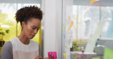 Gülümseyen melez iş kadını beyin fırtınası yapıyor, şeffaf tahtaya notlar yapıştırıyor. Yoğun bir modern ofiste çalışıyor..