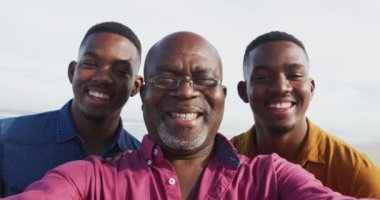 Gülümseyen Afro-Amerikan kıdemli babası ve sahilde selfie çeken ikiz oğulları. Sağlıklı açık hava aile birlikte zaman geçirir..