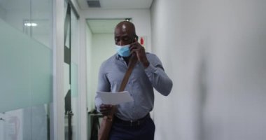 Yüz maskesi takmış Afro-Amerikalı işadamı ofis koridorunda akıllı telefondan konuşan bir belge tutuyor. Modern ofiste çalışan profesyonel iş adamları covid 19 koronavirüs salgını sırasında.