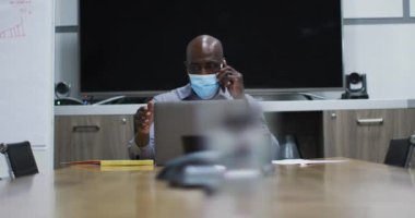 Yüz maskeli Afrikalı Amerikalı iş adamı konferans odasında dizüstü bilgisayarla akıllı telefondan konuşuyor. Modern ofiste çalışan profesyonel iş adamları covid 19 koronavirüs salgını sırasında.