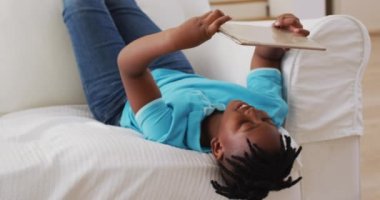 Evdeki kanepede yatarken dijital tablet kullanırken gülümseyen Afrikalı Amerikalı çocuk. Aile, birliktelik ve mutluluk kavramı