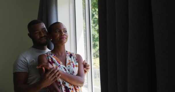 アフリカ系アメリカ人の夫婦が窓から外を眺めながら抱き合っている 夫婦関係のライフスタイルのコンセプト — ストック動画