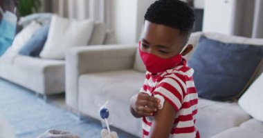 African american doctor wearing face mask gicing a lollipop to a boy after his vaccination at home. vaccination for prevention of coronavirus outbreak concept
