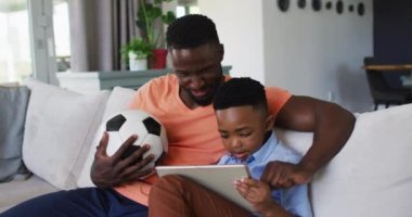 Afro-Amerikan baba ve oğlu evde dijital tabletle spor izlerken birlikte tezahürat yapıyorlar. Aile babası oğul birliktelik ilişkisi kavramı