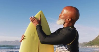 Kıdemli Afro-Amerikan adam sahilde sörf tahtasıyla yürüyor. sağlıklı açık hava eğlence zamanı deniz kenarında.