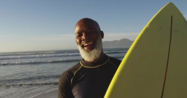 Gülümseyen kıdemli Afrikalı Amerikalı adam sahilde sörf tahtasıyla yürüyor. sağlıklı açık hava eğlence zamanı deniz kenarında.