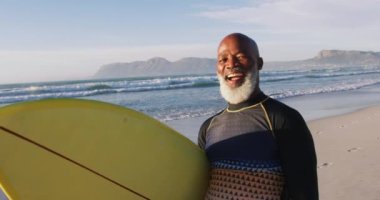 Gülümseyen kıdemli Afrikalı Amerikalı adam sahilde sörf tahtasıyla yürüyor. sağlıklı açık hava eğlence zamanı deniz kenarında 