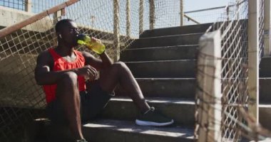Yorgun Afrikalı Amerikalı bir adam oturuyor, su şişesinden içiyor, dışarıda egzersiz yapıyor. fitness, sağlıklı ve aktif yaşam tarzı konsepti.
